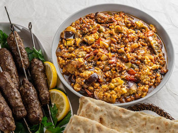 A bowl of colorful lentil stew with vegetables next to grilled kebabs on skewers, lemon slices, parsley, and flatbread on a textured table.