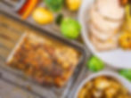 Sliced roast pork with roasted potatoes, carrots, brussels sprouts on a white plate. Colorful veggies and herbs scattered on a marble surface.