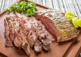 Sliced grilled steak on a wooden board with parsley and lime wedges. The background has a black-and-white striped pattern.