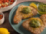 Grilled steaks on a blue plate with green pesto, lemon slices, and a bowl of roasted tomatoes. Rustic wooden table background.
