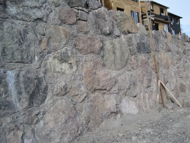 Boulder retaining wall under construction at the West Hills development in Langford, BC, with new home framing visible in the background.