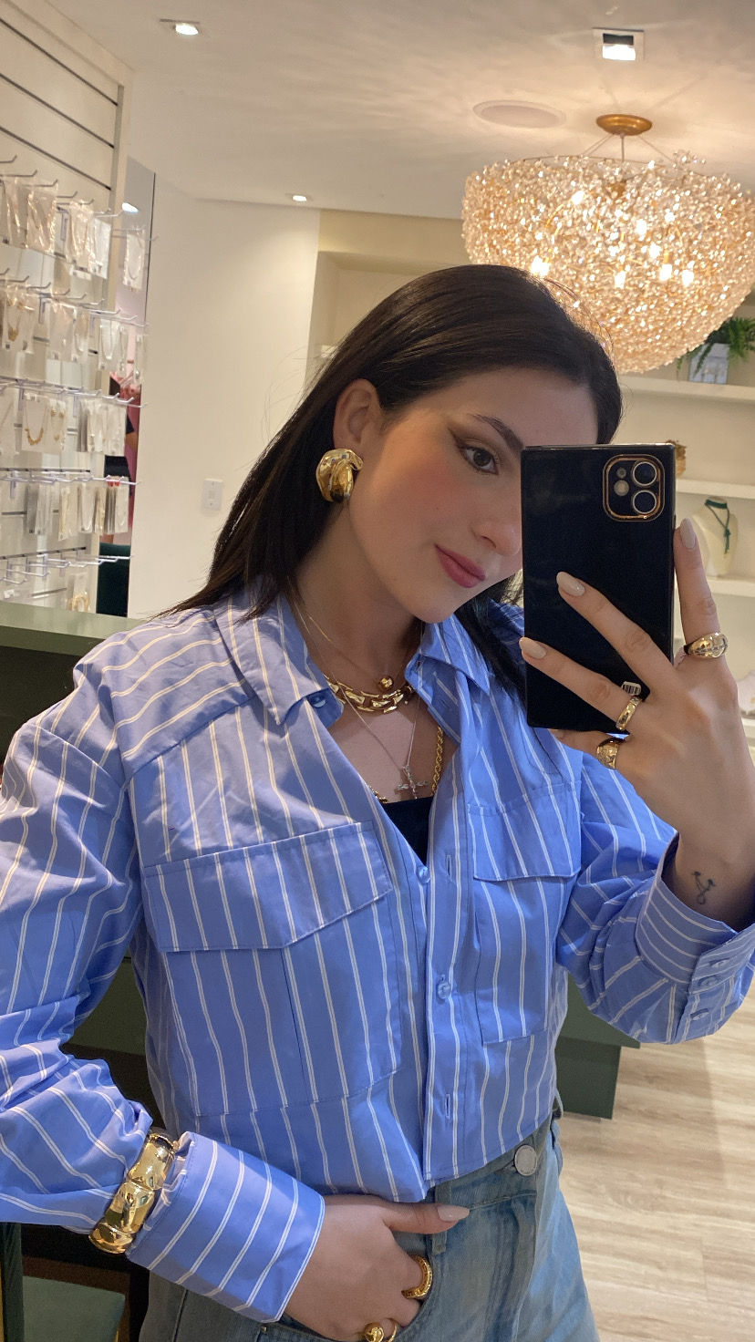 Audiodescrição: Mulher jovem adulta de pele clara e cabelos longos, lisos e pretos. Está em pé, sorrindo e segurando um celular, como se tirasse uma selfie. Seu rosto oval tem nariz fino e lábios cheios. Veste uma camisa azul listrada e está com diversos anéis e colares dourados, além de um brinco dourado grande. O fundo sugere uma loja de roupas ou acessórios.