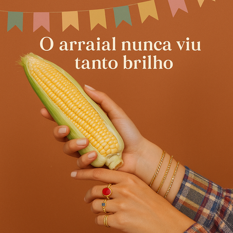 Imagem com fundo laranja terroso e bandeirinhas coloridas de festa junina no topo. Em destaque, duas mãos femininas seguram uma espiga de milho amarelo ainda com a palha parcialmente aberta. As mãos estão adornadas com vários anéis dourados (um deles com pedra vermelha e outro azul) e pulseiras douradas, trazendo um toque sofisticado e brilhante. A pessoa veste uma camisa xadrez, tÃpica de festas juninas. Acima da espiga, lê-se o texto em branco: "O arraial nunca viu tanto brilho", fazendo alusão ao brilho das joias e ao clima festivo.