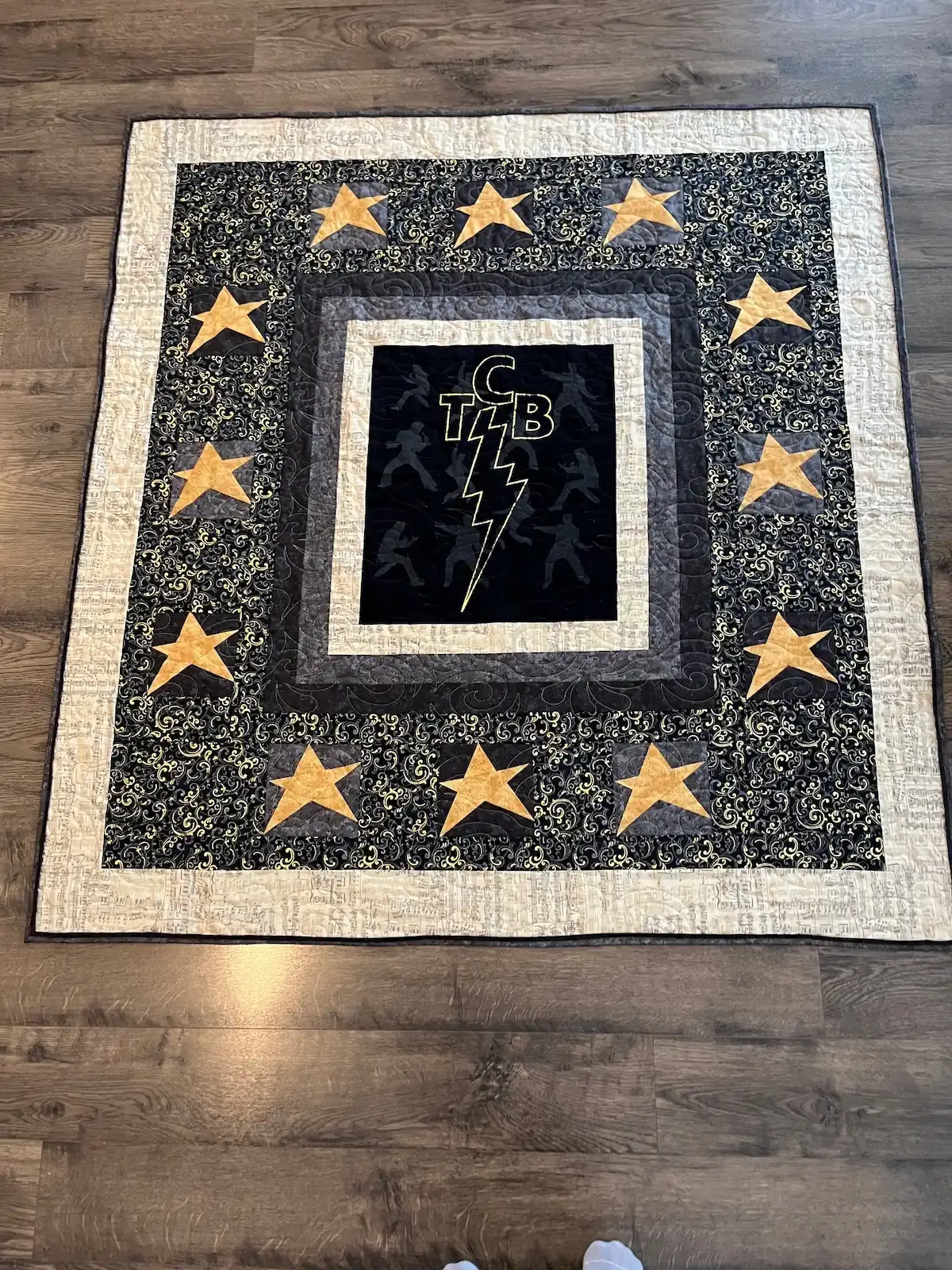 T‑shirt memory quilt featuring TCB lightning‑bolt shirt in the center with black borders and gold stars.