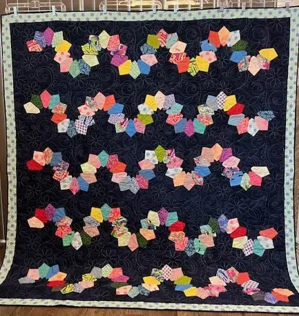 Handmade donated quilt with colorful pentagon‑style flower blocks arranged on a dark navy background with a patterned border.