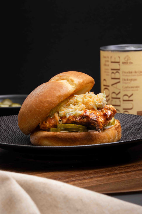 A crispy chicken sandwich on a black plate topped with coleslaw with a visible can of maple syrup in the back.