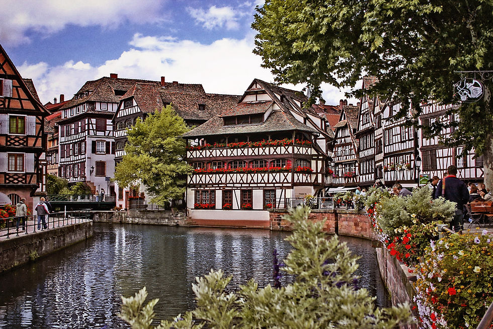 Quaint half-timbered houses line a canal in a European village. Vibrant flowers adorn the scene, and people stroll nearby under a clear sky.