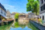 Charming canal with colorful flowerbeds, surrounded by timber-framed houses and a large tree under a clear blue sky. Reflective water surface.