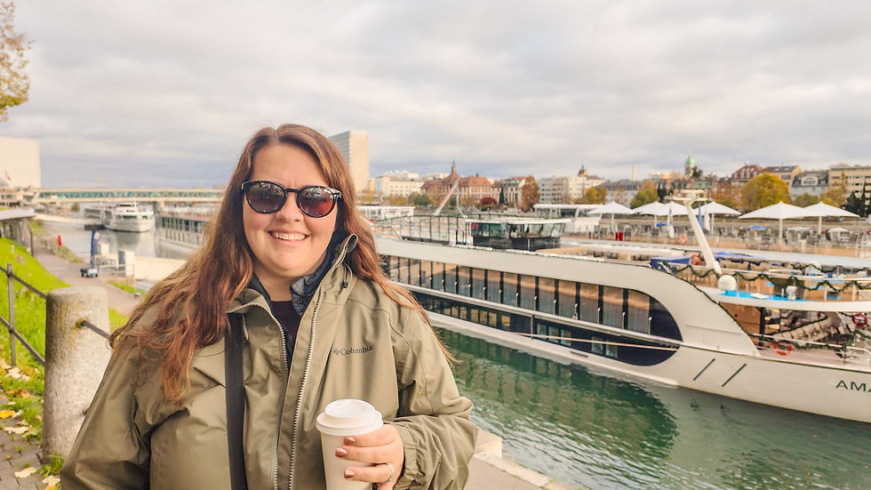 Smiling person in sunglasses holds a coffee cup by a river, with a cruise ship and city buildings in the background under a cloudy sky.