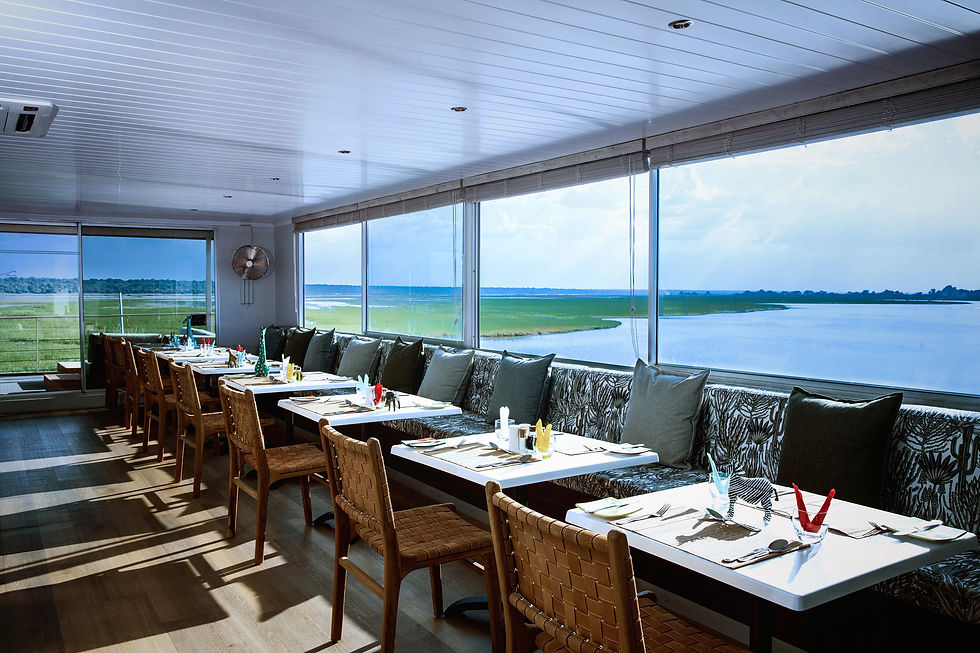 Empty modern riverside dining room with set tables, woven chairs, and wall-to-wall windows overlooking blue water and marshes