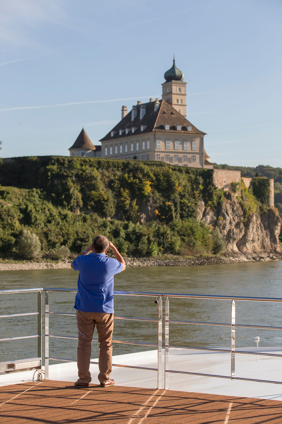 Man in blue shirt photographing a grand castle on a cliff across a river. Clear sky, lush greenery, and tranquil mood.
