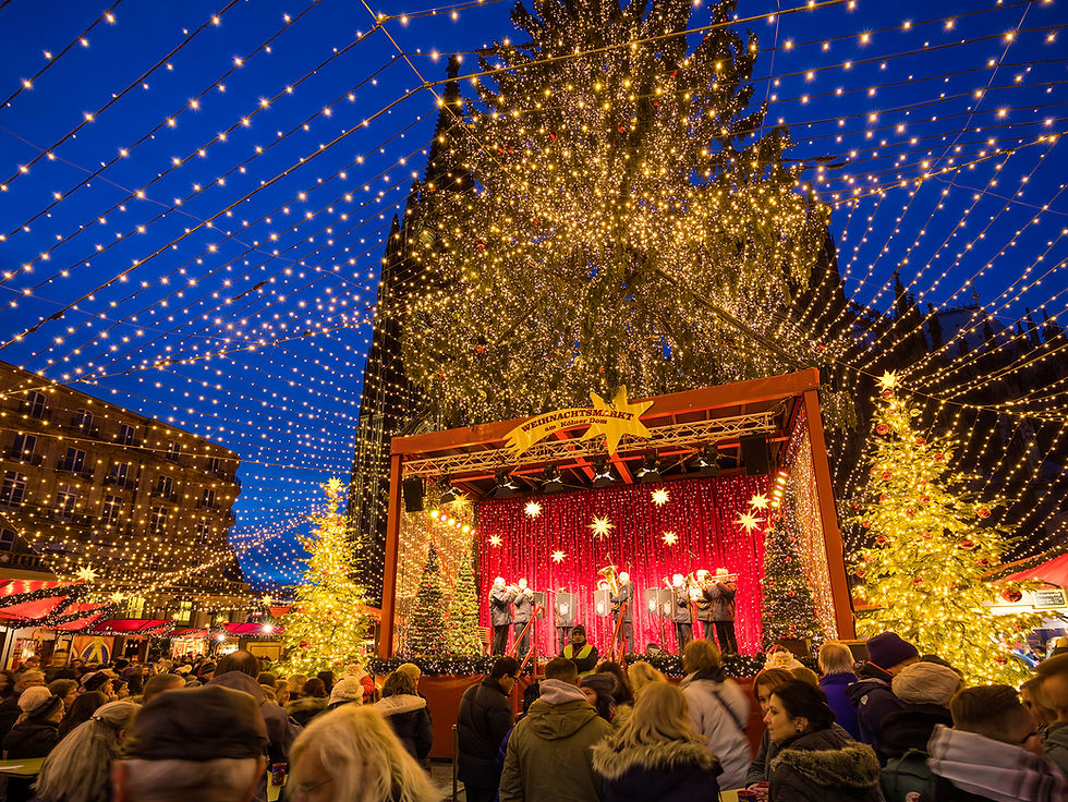 Crowd at a festive Christmas market under string lights. A band plays on stage with red curtains. Decorated trees glow warmly at night.