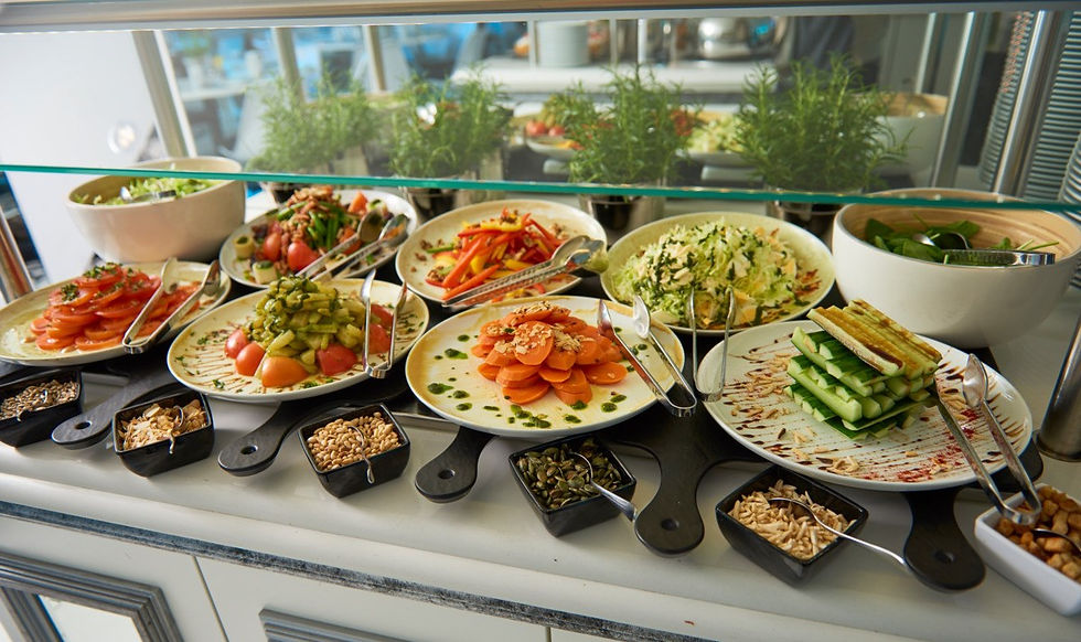 A colorful buffet featuring plates of fresh vegetables and salads with tongs, small bowls of seeds, and herbs in the background.