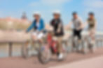 Family of four biking on a boardwalk by a river. They wear helmets and casual clothing. Cityscape in the background under a clear sky.