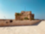 Historic fortress with round towers under a clear blue sky. Stone path leads towards it, lined with palm trees. People walk leisurely.