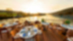 Dining tables with blue-patterned plates on a wooden deck overlooking a serene river at sunset, surrounded by trees and hills.