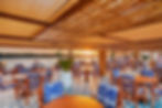 Outdoor terrace with wooden tables and blue patterned chairs, overlooking a river at sunset. Warm lighting, leafy plants, relaxed ambiance.