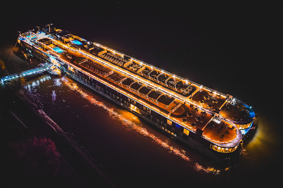 Aerial view of a large, illuminated cruise ship at night on a dark river. Bright lights outline the decks, creating a festive ambiance.