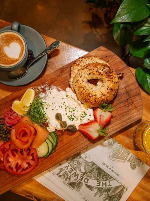 overhead photo of a bagel platter with smoked salmon and freshly chopped vegetables next to a cappuccino.