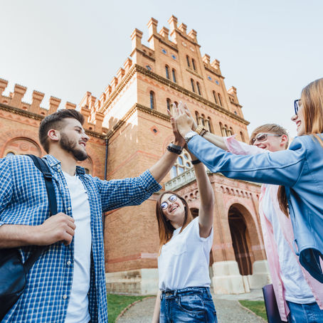 Group of four young students high-fiving outside a historic brick building, showing happiness and teamwork, with clear blue sky above.