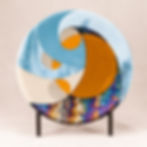 A circular abstract glass sculpture featuring swirls of rust, blue, cream, and black irid glass interacting