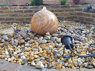 A smooth, spherical stone water feature sits on a bed of multicolored pebbles