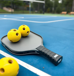 a gray pickleball paddle with 3 pickleballs on top of it sitting diagonally on a blue cour