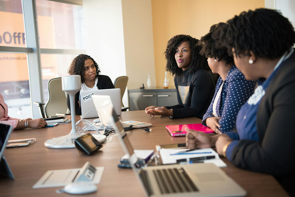 Five women in a meeting room, engaged in discussion. Laptops and notepads on the table. Bright window and neutral walls in the background.