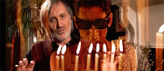 Two men, one with long hair and another wearing sunglasses, superimposed over flickering candles in a warm, dimly lit room.