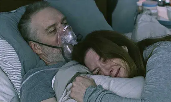 A man in a hospital bed with an oxygen mask, a woman lies beside him, hugging. Dimly lit room, somber mood. No visible text.