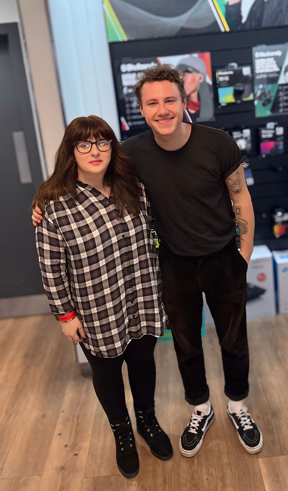 Two people stand smiling in a store. One wears a black t-shirt, the other a black and white checkered dress. Background displays electronics.