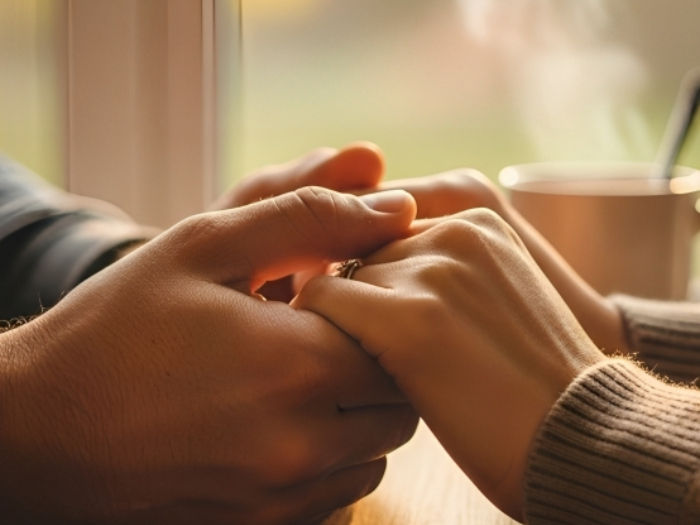 Pair of hands holding each other warmly by a window. Soft light, brown tones, steaming cup in background. Mood is cozy and affectionate.