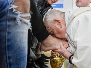 Elderly man in white robe kisses foot of seated person, holding a golden container. Background shows people in casual attire. Emotional scene.