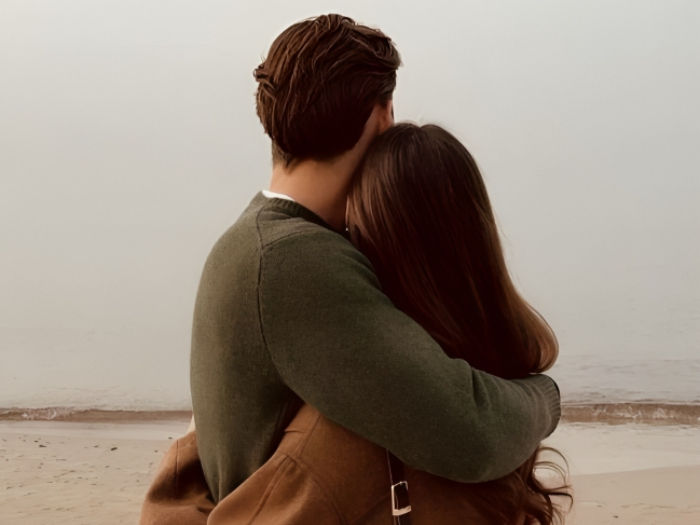 Two people embrace on a foggy beach, facing the ocean. The mood is peaceful. The scene features earth-toned clothing and a serene atmosphere.