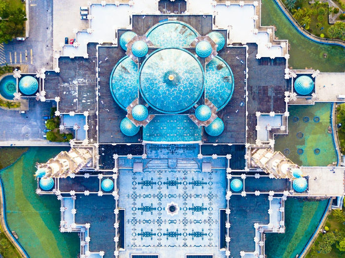 Aerial view of a mosque with intricate turquoise domes and ornate patterns, surrounded by lush greenery and serene water, creating a tranquil mood.