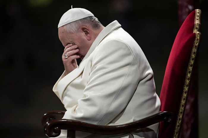 A man in white attire sits on a red chair with head resting on hand, appearing contemplative, against a dark background. No visible text.