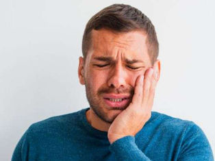 A man in a blue sweater holds his cheek, wincing in pain against a plain background, conveying discomfort.