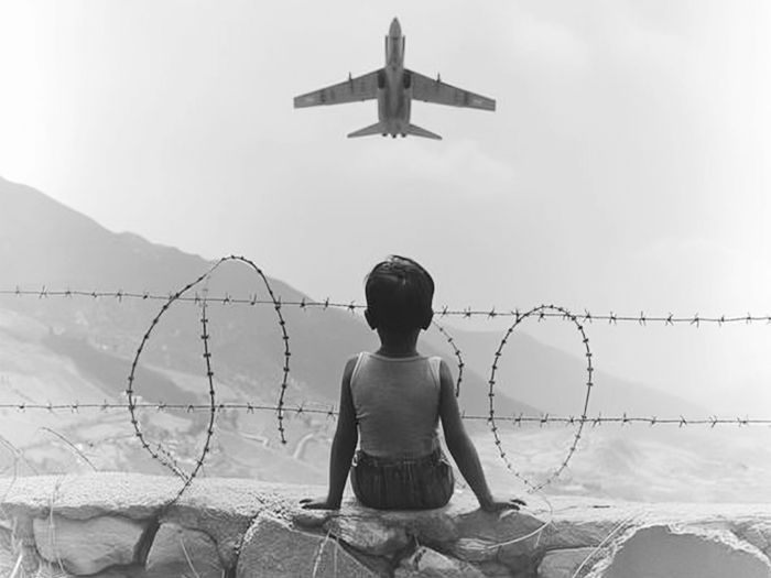 A child in a tank top sits on a wall, watching a jet fly overhead. Barbed wire and mountains in the background create a somber mood.