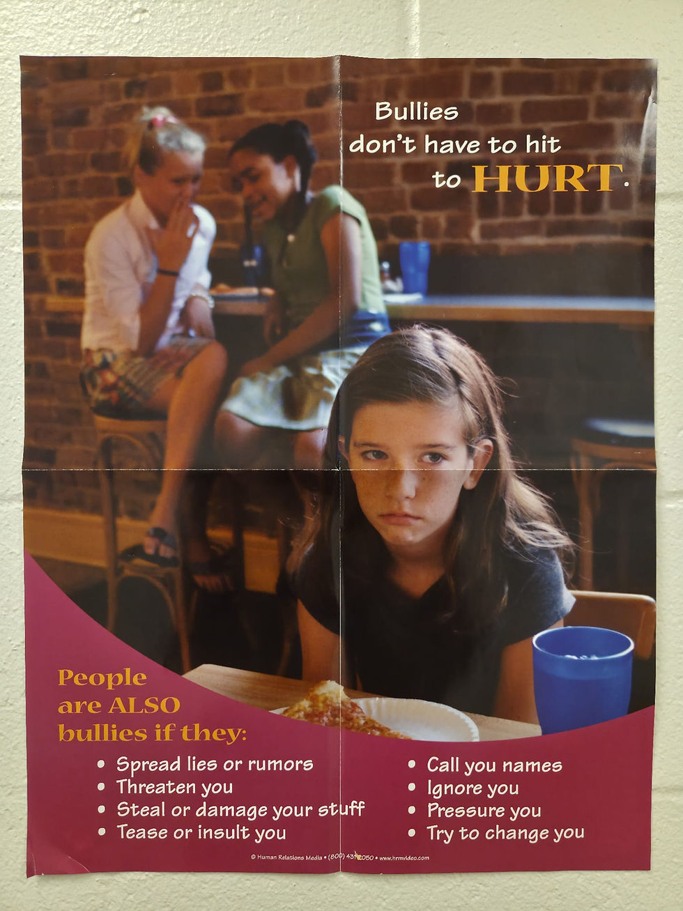 Poster showing a sad girl at a table, two girls gossip in the background. Text: "Bullies don't have to hit to hurt." Bullying actions listed.