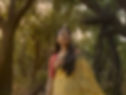 A woman in a yellow sari with polka dots stands in a serene forest, smiling slightly, surrounded by tall trees and dappled sunlight.