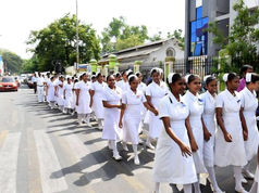 A group of nurses in white uniforms walk in a line on a street, bordered by trees and buildings with blue accents. It's a sunny day.
