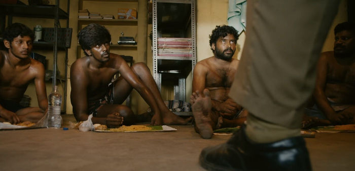 Shirtless men sit on the floor with meals; a person in police uniform stands in front. Shelves and muted colors set a tense mood.