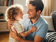 A father and child smiling at each other in a cozy living room. The child wears a striped shirt, and light fills the warm, inviting space.
