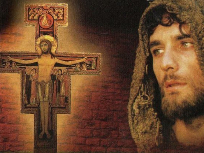 St Francis of Assisi in a hood looks towards a decorated crucifix against a brick background. Warm tones and a contemplative mood dominate.