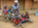 A woman in a headscarf sits with five children on a dirt floor outside a thatched house. The kids wear colorful clothes, appearing calm.