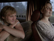 Two people in a vehicle, a woman driving and a man leaning forward, smiling. The interior is dimly lit with blurred trees outside.