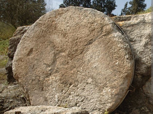 Giant stone disc standing upright in a rocky outdoor setting, surrounded by trees. Earthy tones dominate the scene.