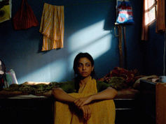 Woman sitting pensively in a dim blue room, sunlight streaming in. She wears a yellow sari. Clothes hang behind her, enhancing a reflective mood.
