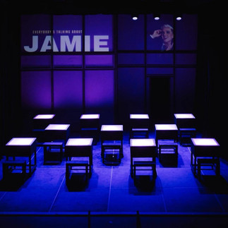 The opening stage of JAMIE, 11 desk that have lights inside sit on the stage evenly spaced, their is a projection on the back wall of the show title, the mood lighting is blue