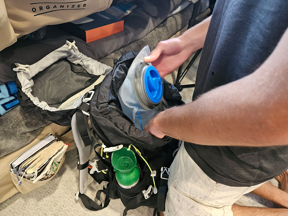 Person with grey T-shirt and khaki shorts from chest down packing a full camel pack into a hiking/backpacking pack